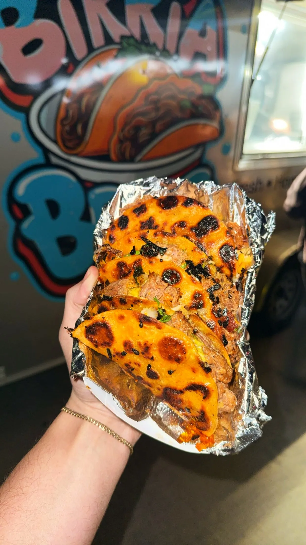 A hand holding a foil tray of crispy, cheesy birria tacos in front of the Birria Bros truck