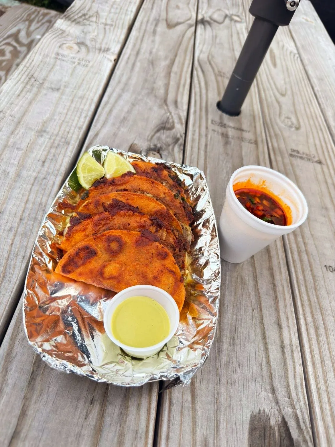 Crispy birria tacos in foil with a cup of consomé and salsa verde on a picnic table